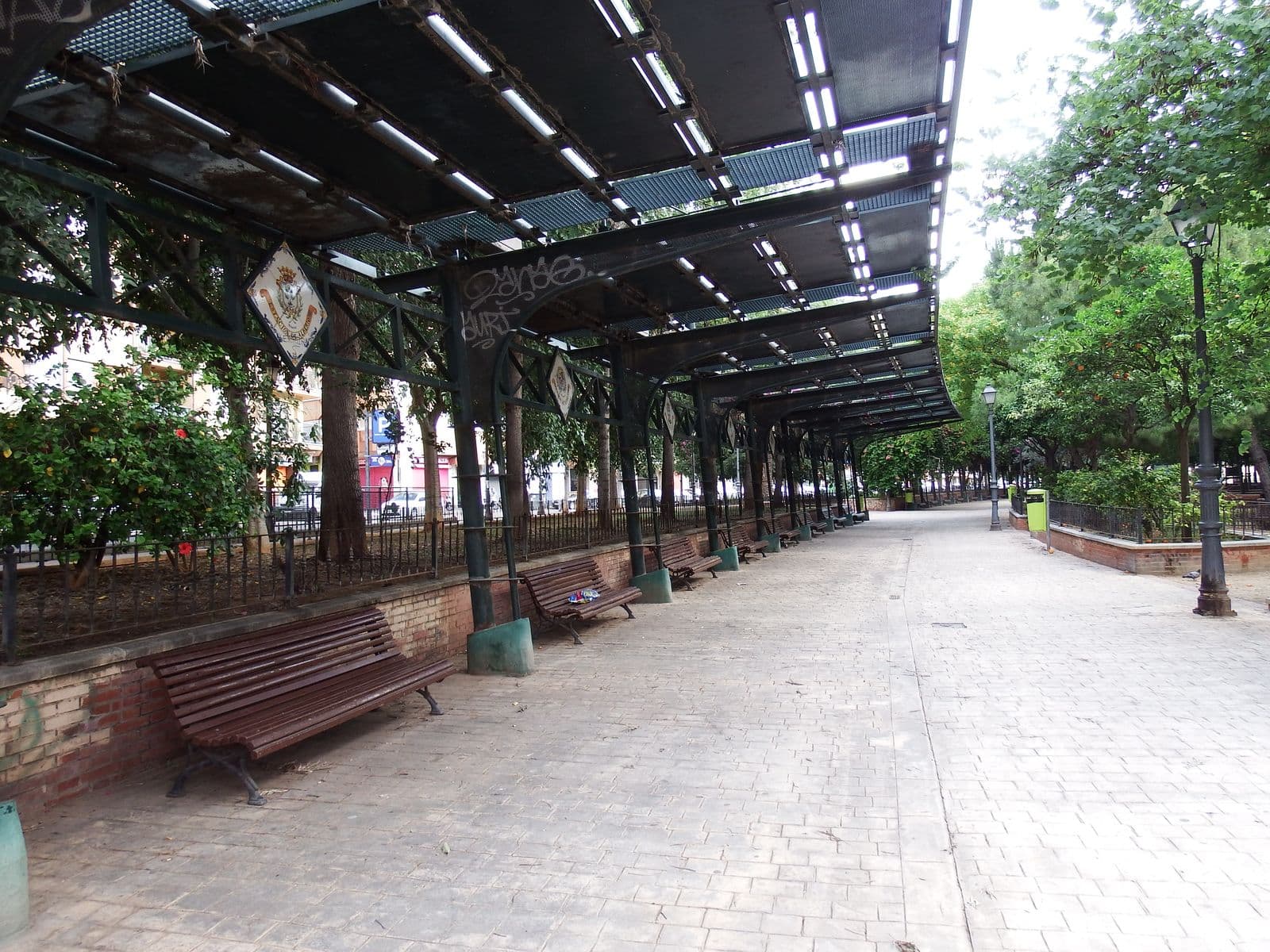 Le jardin de l’Estacioneta à Valencia : allée pavée sous les anciennes marquises métalliques de la gare, ornées de blasons en céramique, bancs et arbres