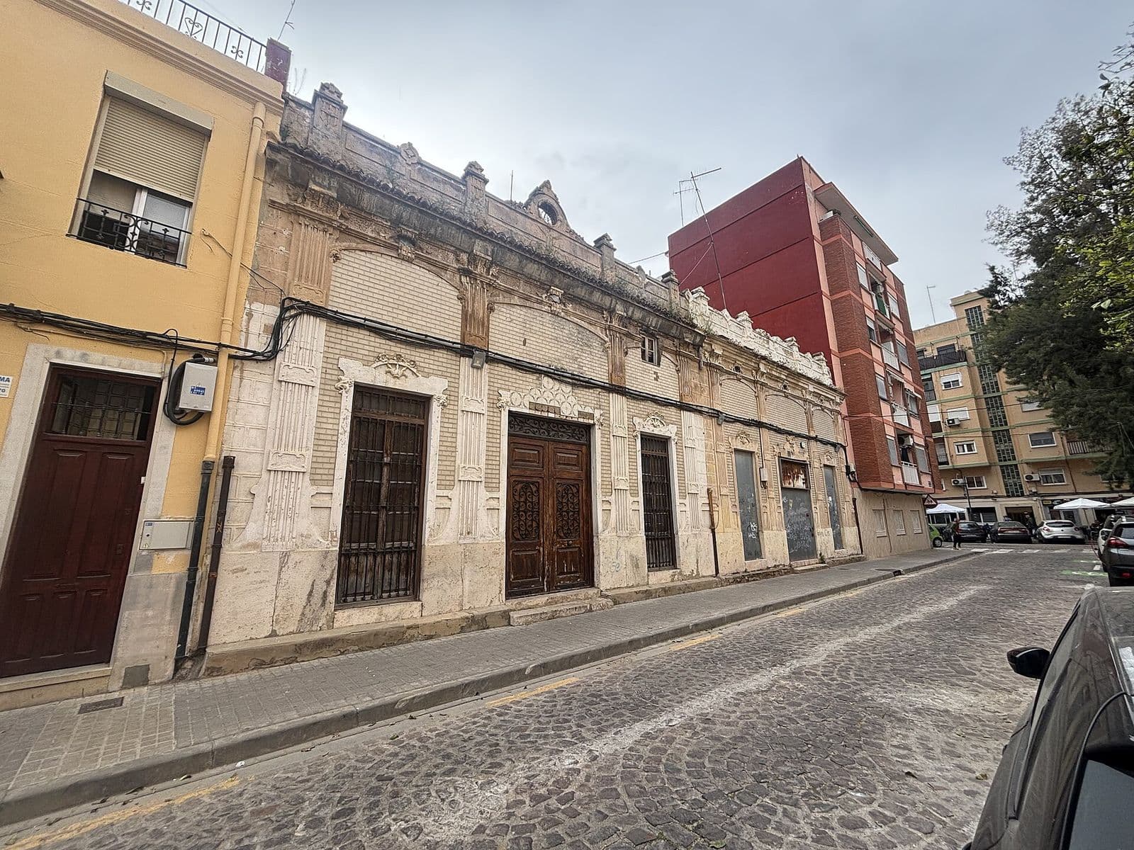 Maison ancienne à façade ornée et portes en bois sculpté dans une rue pavée de Nazaret, à côté d’un immeuble d’habitation en brique rouge (Valencia)