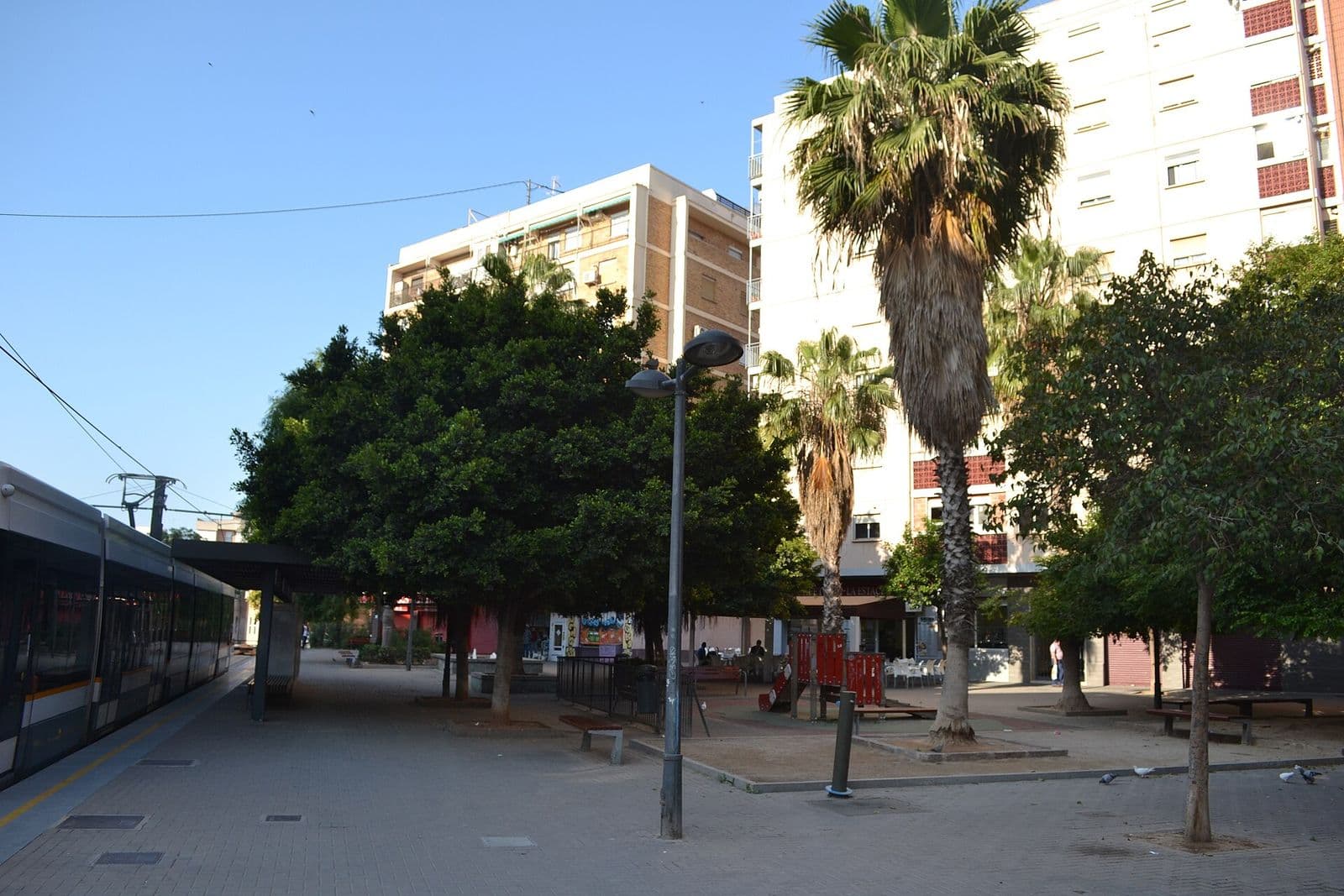 La plaça del Tossal del Rei à Torrefiel : une rame du tram L6 à gauche, des palmiers, des arbres et des immeubles d’habitation (Valencia)